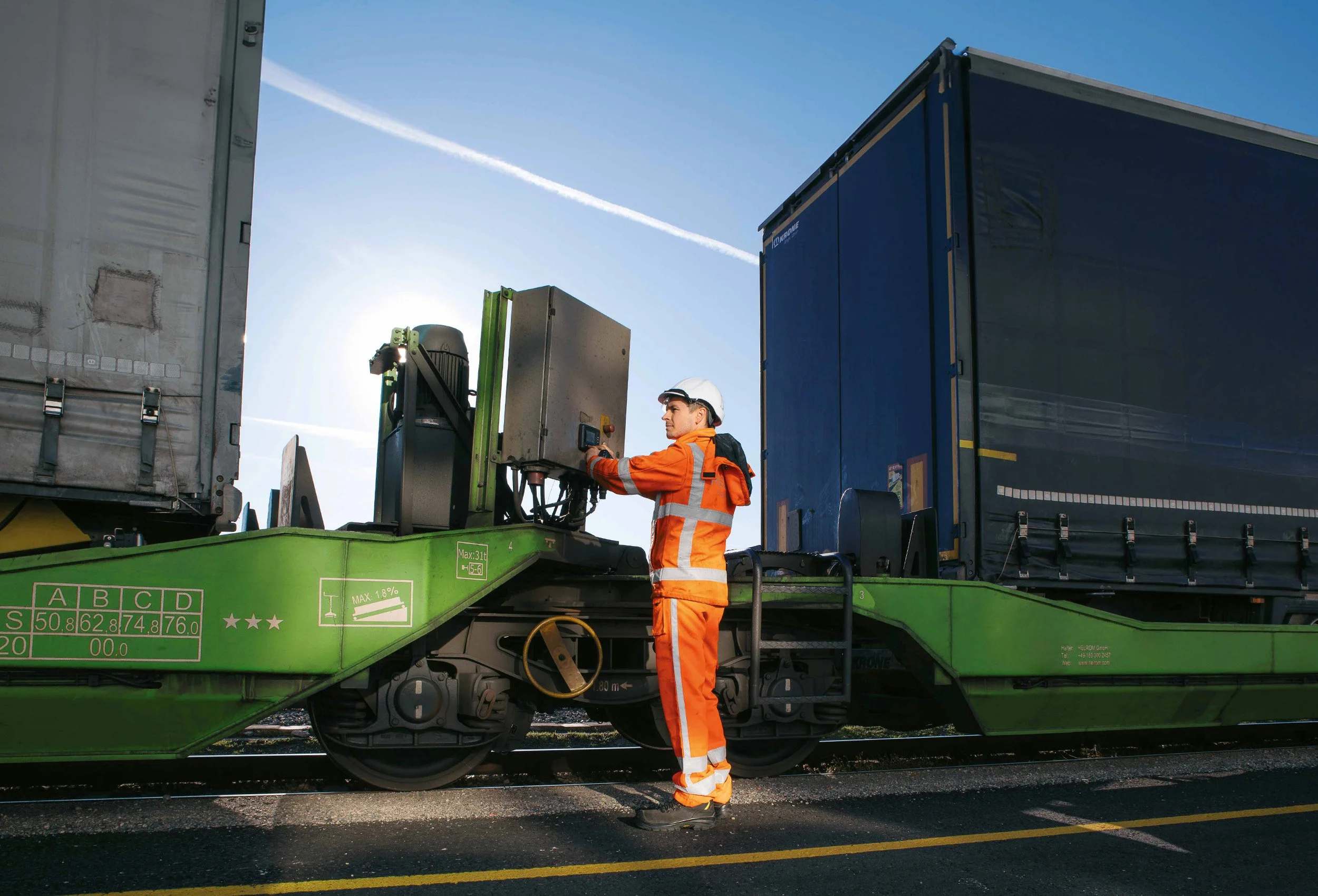 Eine Person in orangefarbener Warnschutzkleidung bedient bei tief stehender Sonne den Schaltkasten am grünen Trailerwagen, daneben ein beladener Sattelauflieger.