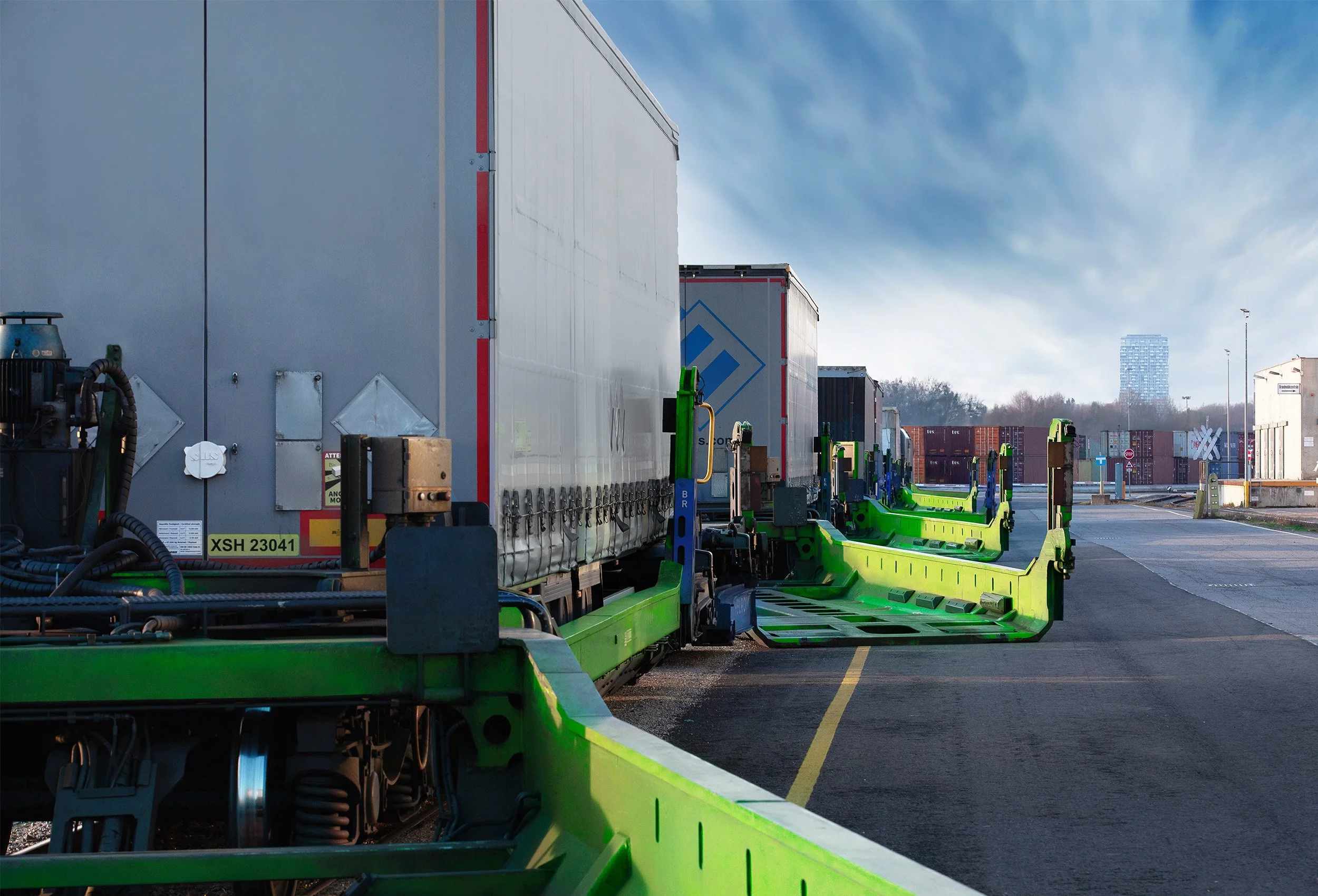 Eine Reihe grüner Trailerwagen bei tief stehender Sonne, mit beladenen Sattelaufliegern und offenen, leeren Ladetaschen entlang der asphaltierten Fläche.