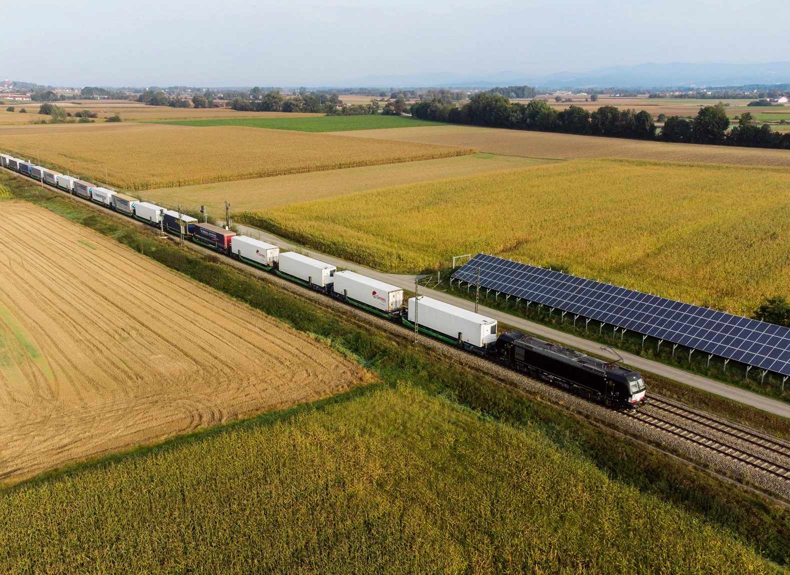 Aerial view: a fully loaded trailer train travelling through farmland, passing a solar array.