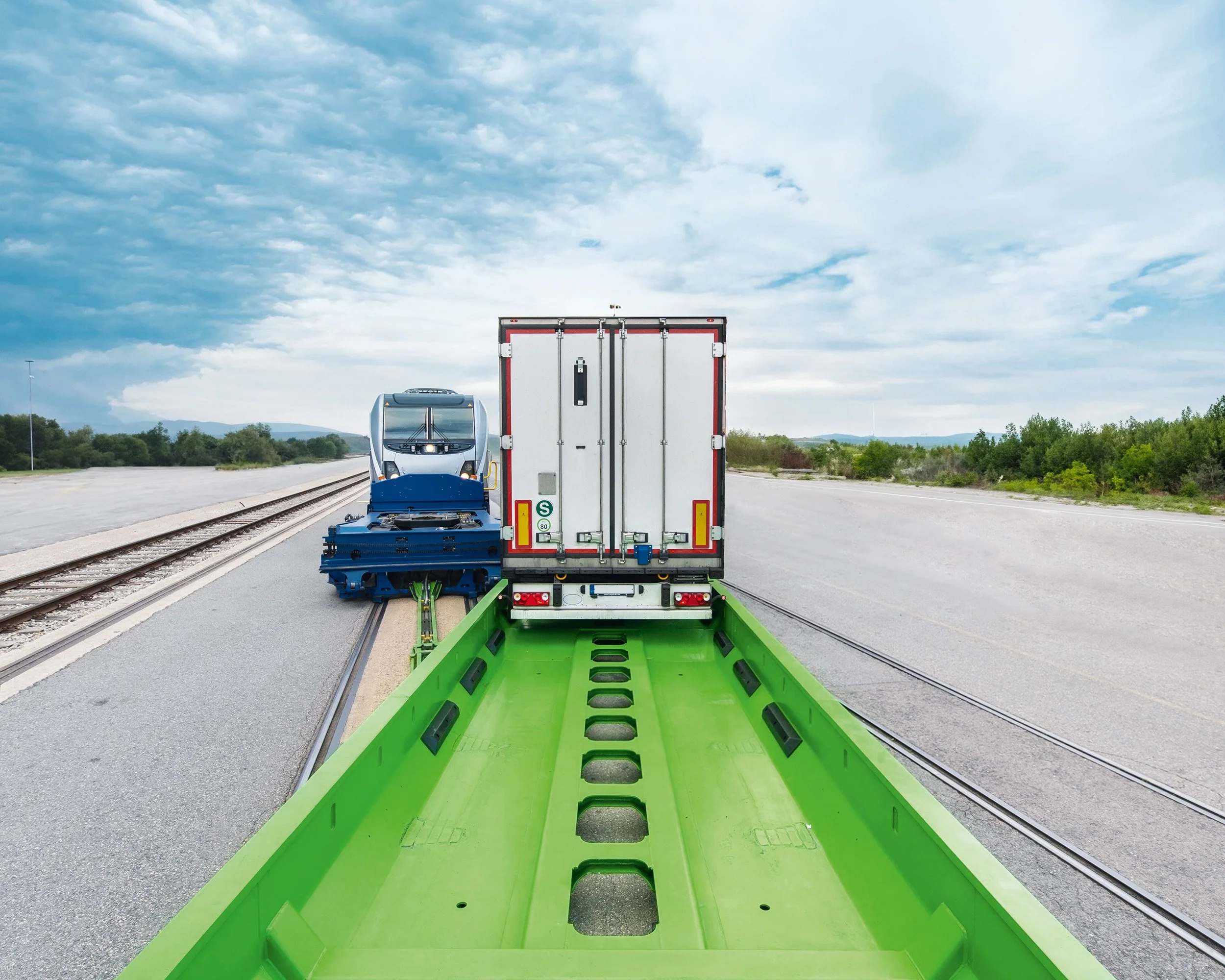 Looking along the open green loading pocket of a trailer wagon: the empty trough in the foreground, a loaded semi-trailer beyond it, the locomotive at the head of the train.
